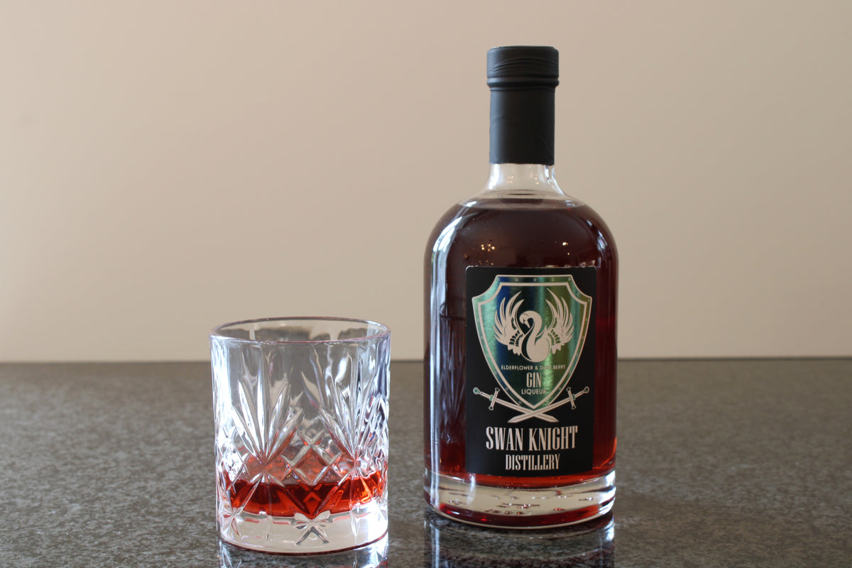 A bottle of Swan Knight Distillery Elderflower and Dark Berry Gin Liqueur on a dark countertop with a filled glass beside it.