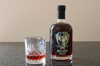 A bottle of Swan Knight Distillery Elderflower and Dark Berry Gin Liqueur on a dark countertop with a filled glass beside it.
