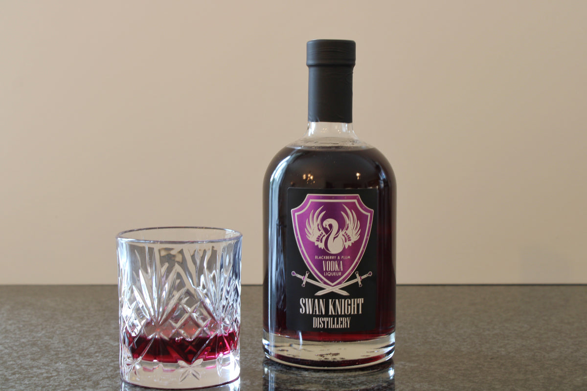 A bottle of Swan Knight Distillery Blackberry and Plum Vodka Liqueur on a dark countertop with a filled glass beside it.